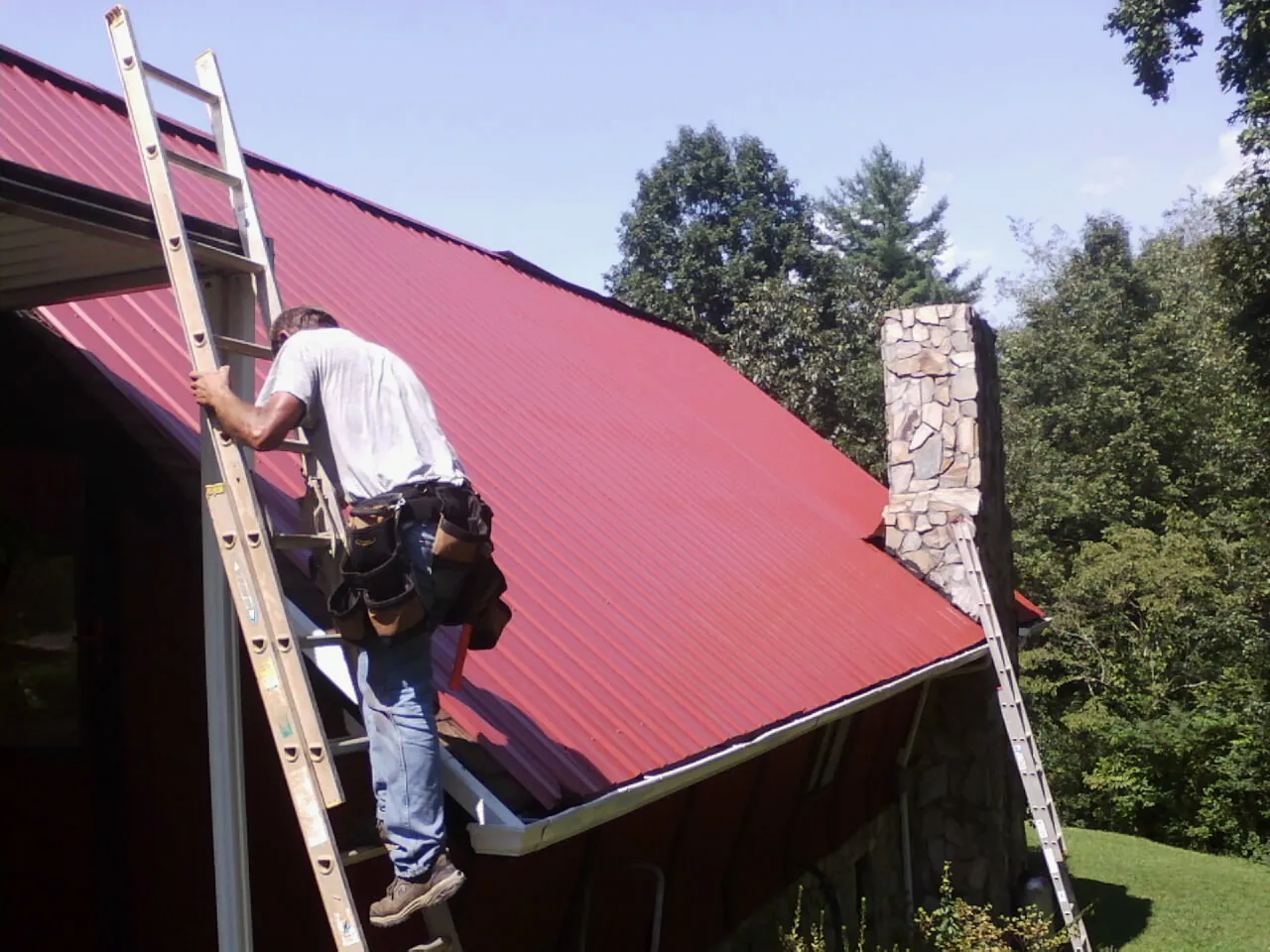 Roofer on a ladder beside a red metal roof and stone chimney — Soffit & Fascia Repair by Roofing Brothers in Elliottsburg